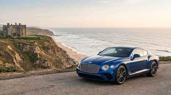 Sequin Blue Bentley Continental GT parked on a scenic coastal road with a manor house and ocean in the background, optimized to 37 KB.